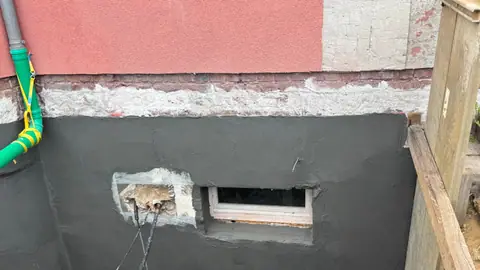 Freigelegte Kellerwand von außen mit frischer dunkler Abdichtung rund um ein Kellerfenster.