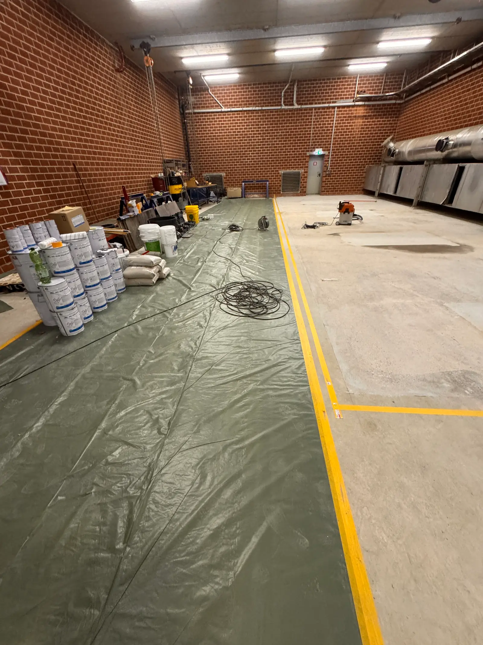 Industriehalle vor der Bodensanierung: alter Betonboden mit Flecken und Ausbesserungen, ein Teil ist mit Folie abgedeckt.
