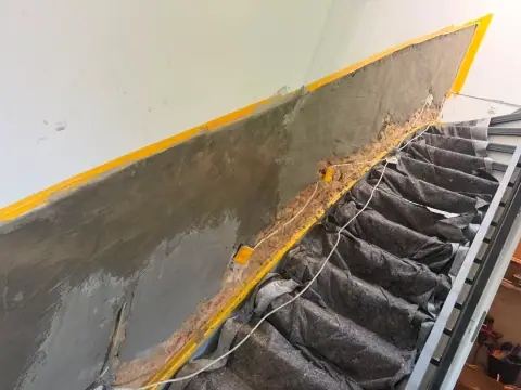 Treppenhauswand mit frisch aufgetragenem Sanierputz im unteren Bereich, abgeklebt zur gestrichenen Wand darüber.